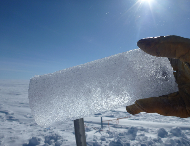 A 25-cm long ice core is held in front of the camera on a sunny day. The background is an endless snow-covered flat landscape and a bright blue sky.