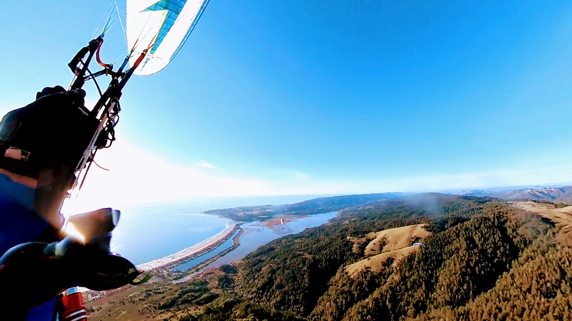 First person view while paragliding during a sunny day with blue skies. The pilot's left hand with a glove can be seen pulling the left break with lines going up to the white, left wing tip above. The pilot is in a left turn high above the mountain side with open patched of grass among the tree. Two other paragliders further down can be seen in the distance. Down below, to the left, there is a long ocean beach slowly curving up towards a point in the horizon. Inside the beach, there is a lagoon. Part of the mountain ridge can be seen to the right.